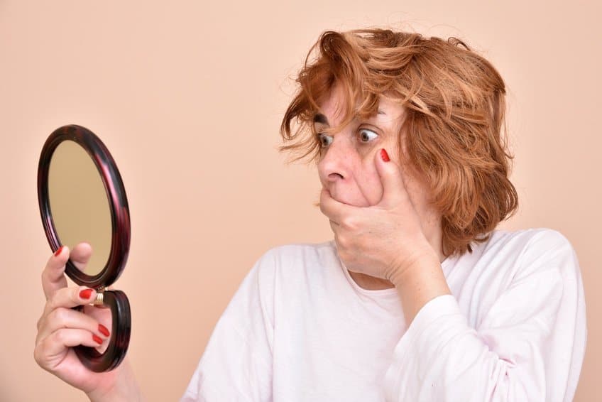 Shocked and surprised middle aged woman looking at her skin and unkempt hair in the mirror Frau die in einen Handspiegel schaut und sich die Hand vor den Mund hält weil sie erschreckt ist